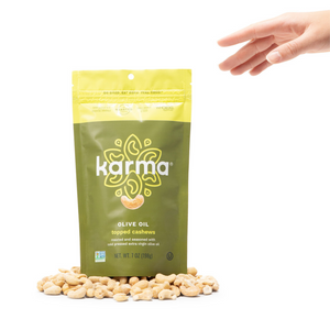 front panel of karma nuts olive oil topped cashews on white background with pile of cashews at bottom of bag and hand reaching for bag 